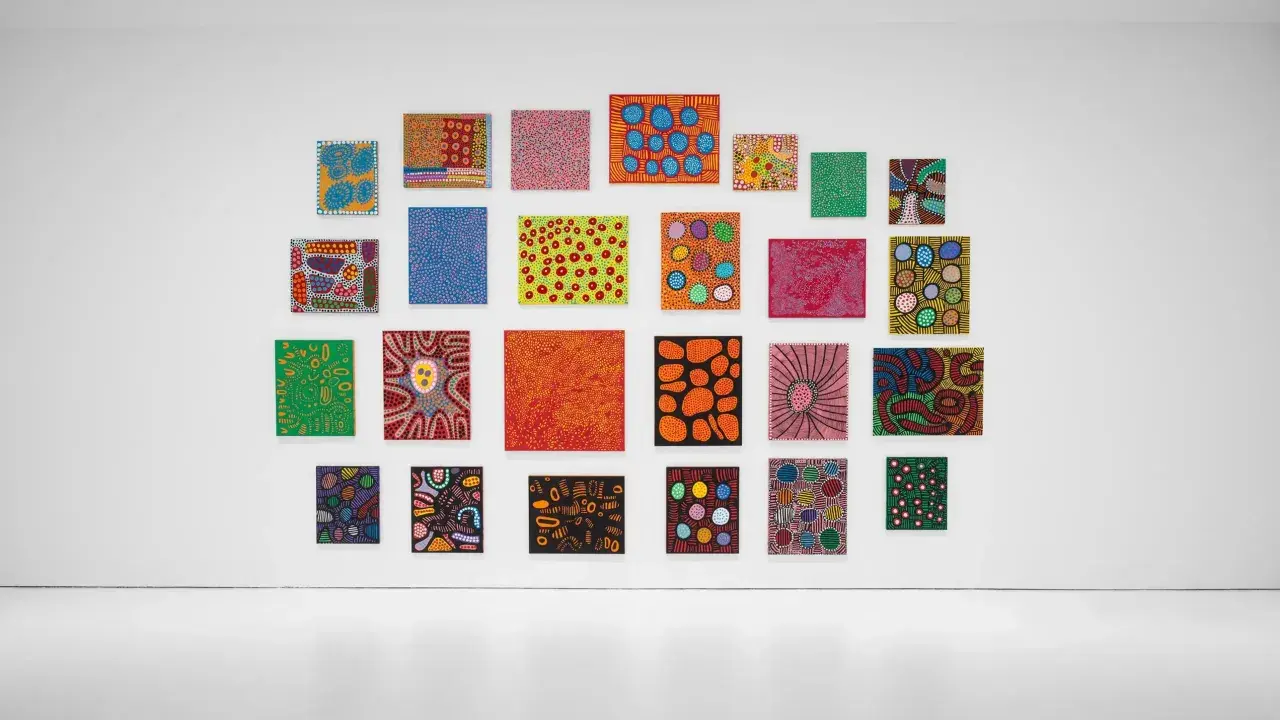 A vibrant collection of paintings by top visual artists, featuring bold colors and abstract patterns, adorns a white gallery wall.
