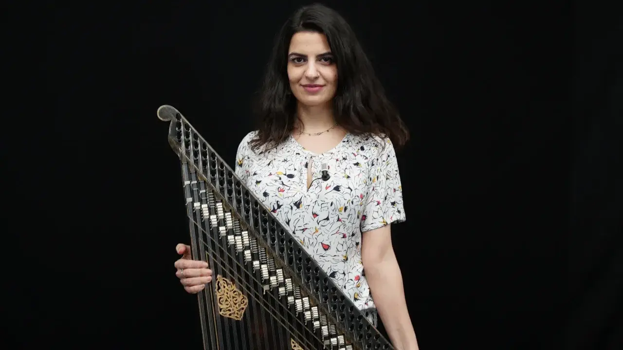 Une musicienne tient un kanoun, instrument de musique à cordes pincées, devant un fond noir.