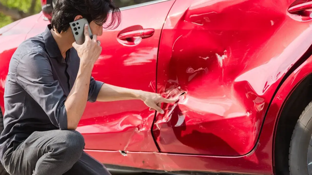 Hombre llama por teléfono señalando el grave golpe en la puerta de su coche rojo.