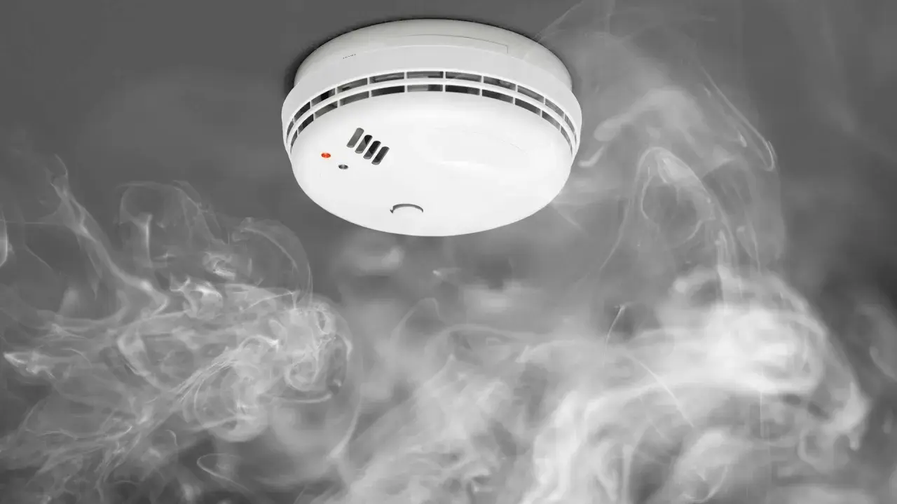 Biały czujnik dymu na suficie, otoczony kłębami dymu. Widać czerwoną diodę sygnalizującą alarm.