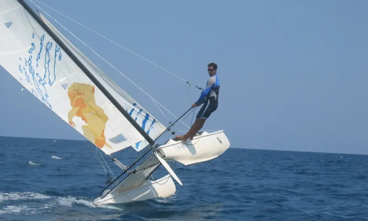 Un homme en trapeze sur un catamaran navigue sur une mer bleue sous un ciel clair. La voile arbore un dessin original.