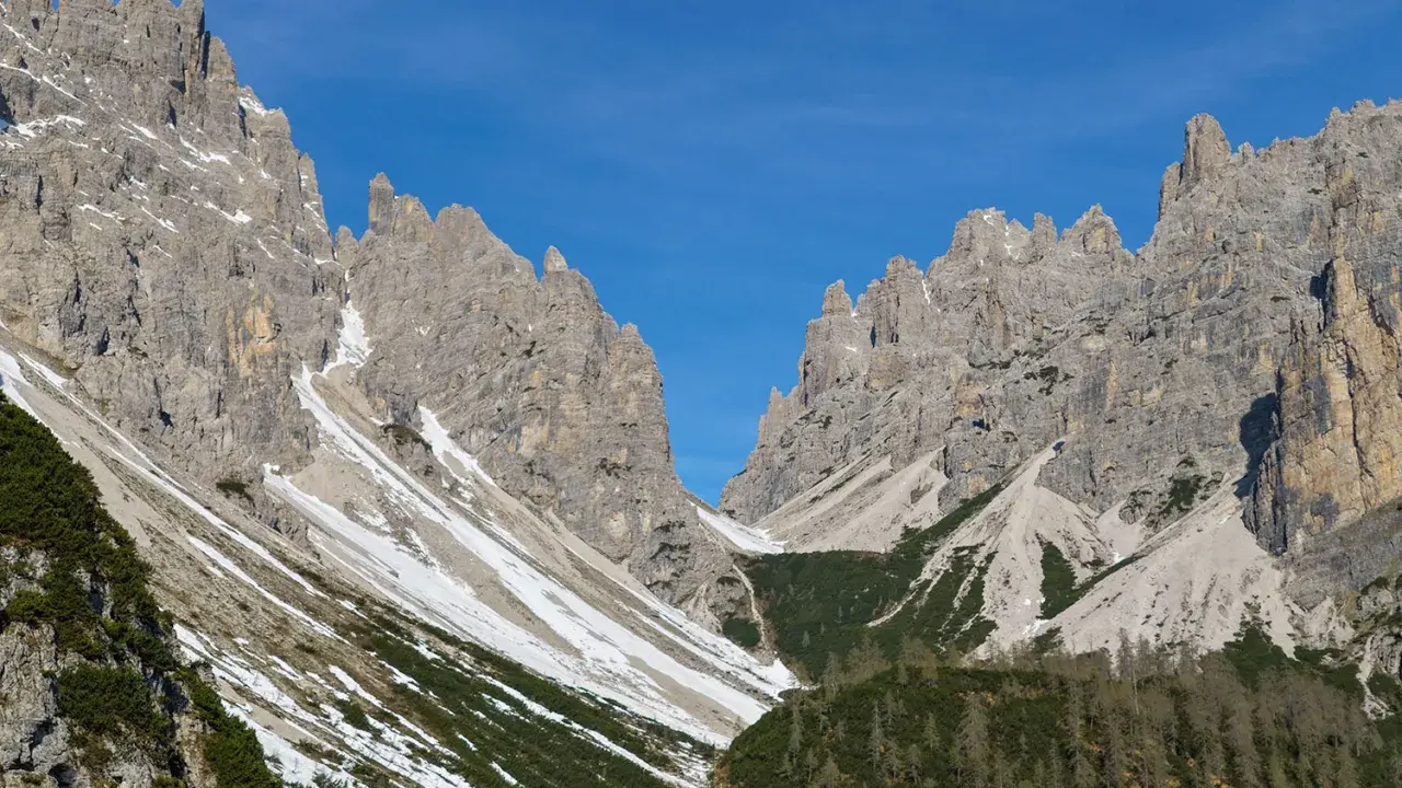 Vette rocciose imponenti sotto un cielo azzurro. Un sentiero scende verso il rifugio Giaf, tra pini e chiazze di neve.