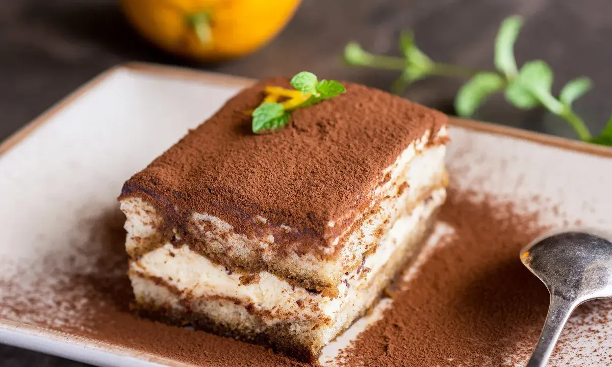 Pyszne tiramisu na biszkopcie, posypane kakao, z miętą i skórką pomarańczy. Obok łyżeczka i pomarańcza.