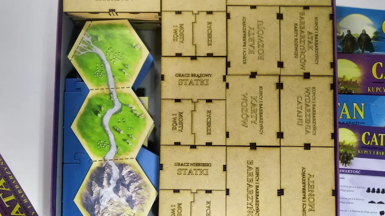 W pudełku dodatku do gry Catan leżą heksagonalne pola z rzeką, góry i lasy, a także liczne karty i żetony z napisami "Kupcy i Barbarzyńcy".