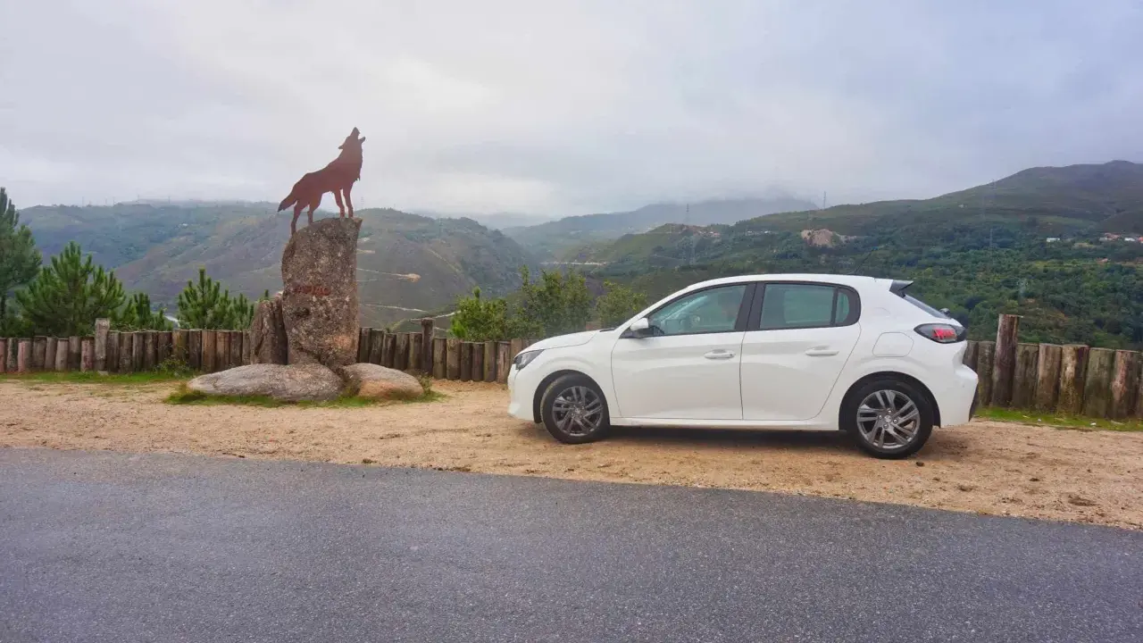 Ein weißer Kleinwagen parkt neben einer Wolfsstatue auf einem Felsen mit Blick auf eine bergige Landschaft. Tolle Carjet Erfahrungen für diese Reise!