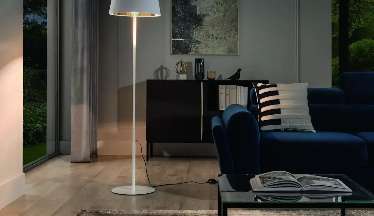 Biała, minimalistyczna lampa stojąca oświetla przytulny salon z granatową kanapą i szklanym stolikiem.