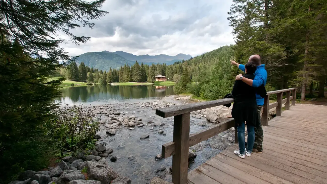 Coppia ammira il lago dei caprioli, con montagne sullo sfondo e una casa tra gli alberi.