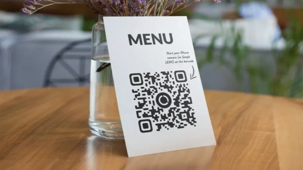 Un código QR de restaurante en una mesa de madera, junto a un jarrón con flores secas. Escanea para ver el menú.