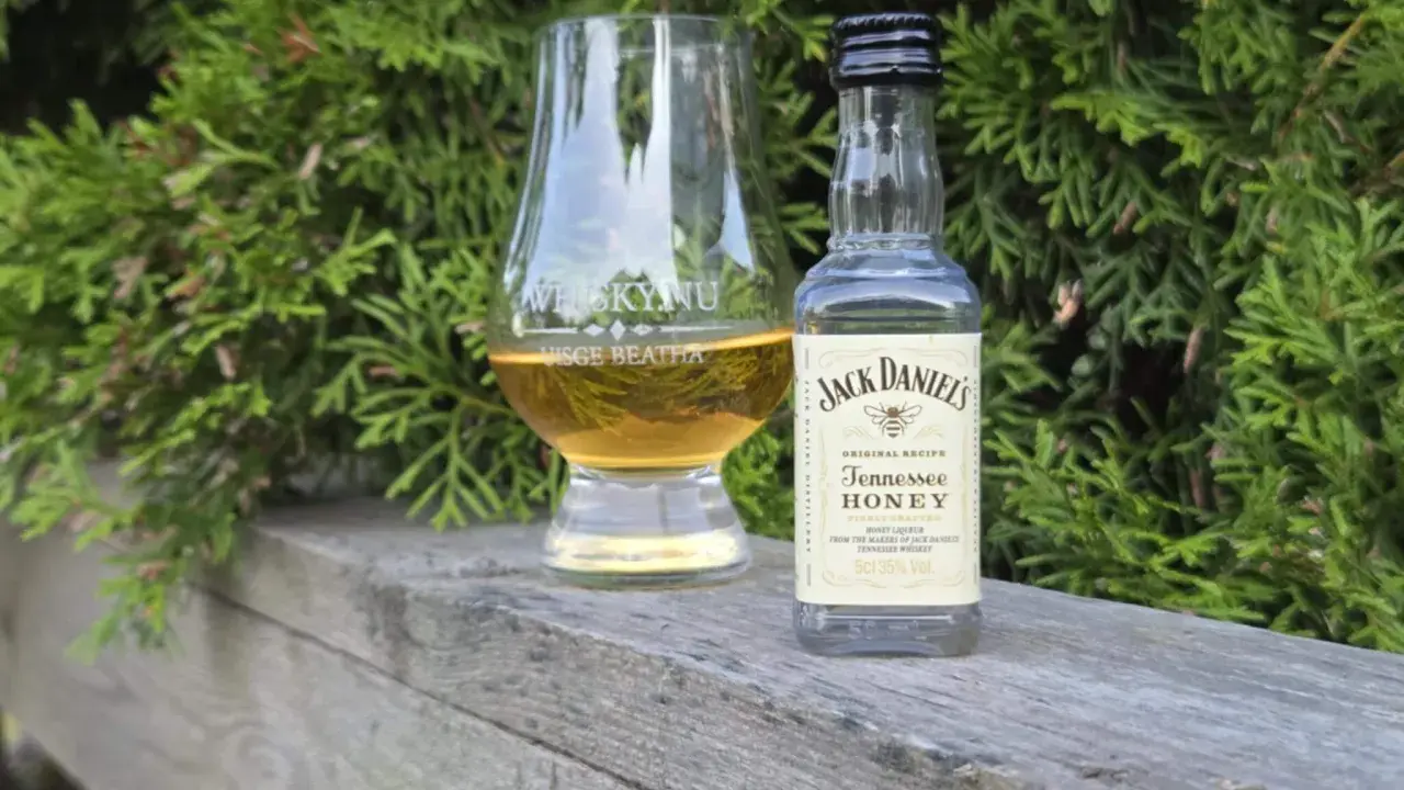 En liten flaska Jack Daniels Honey och ett whiskyglas står på en träbänk, omgivna av grönska.