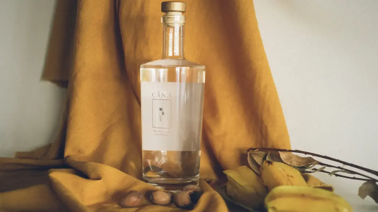 Butelka Cachaça Cãna, brazylijskiego alkoholu z trzciny cukrowej, otoczona owocami i suchymi liśćmi. Cachaca co to? To brazylijski skarb!