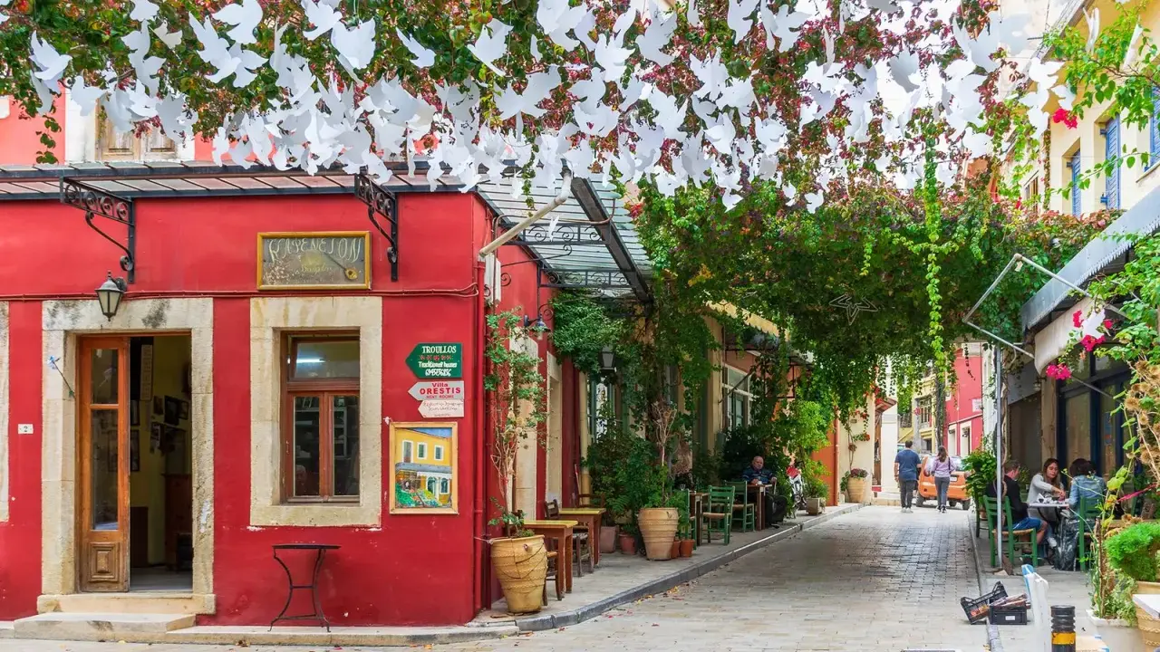 Ein rotes Gebäude mit weißen Tauben-Girlanden über einer Gasse in Chania, Kreta. Tische und Stühle laden zum Verweilen ein.