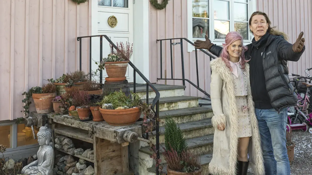 Melinda Jacobs och E-Type poserar glatt framför ett rosa hus med många krukor och en Buddha-staty.