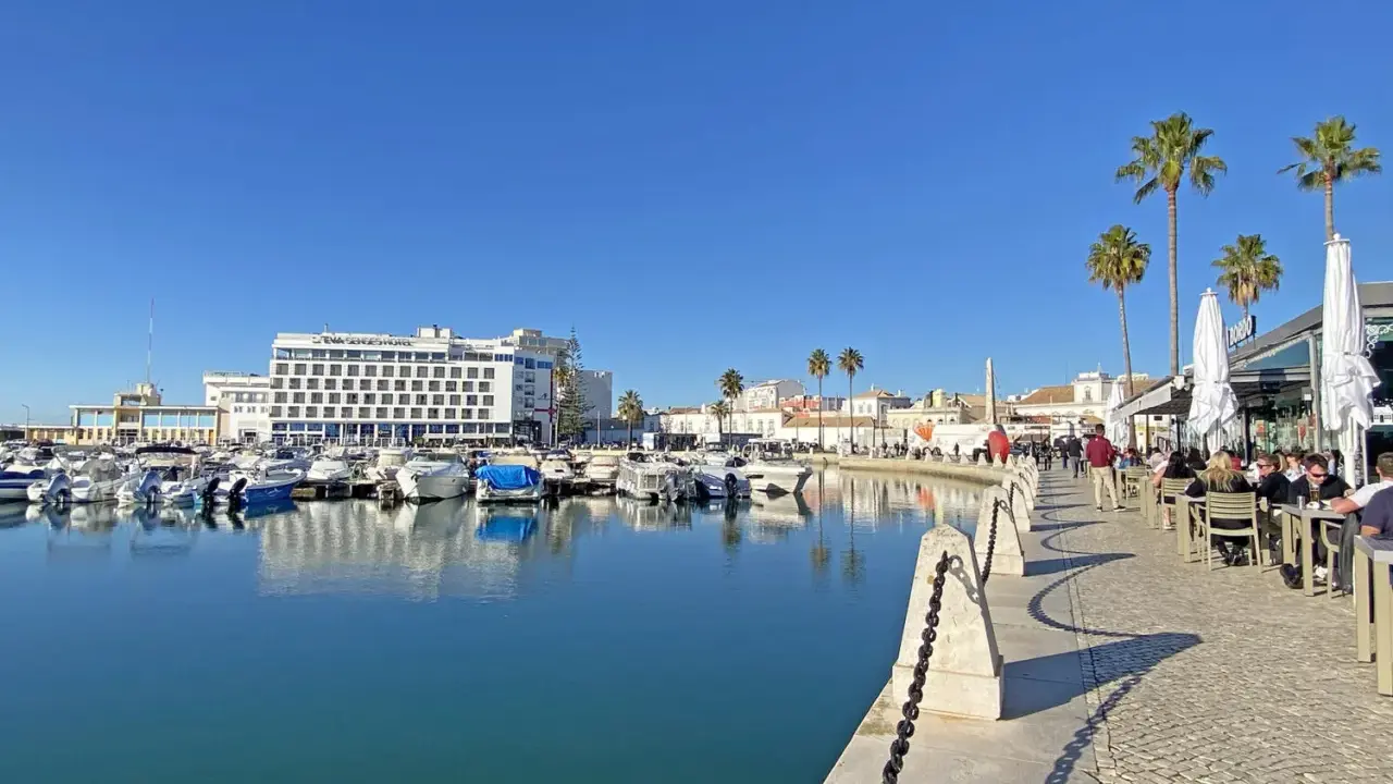 Boote im Hafen von Faro, Portugal, spiegeln sich im ruhigen Wasser. Palmen säumen die Promenade, wo Menschen in Cafés sitzen.