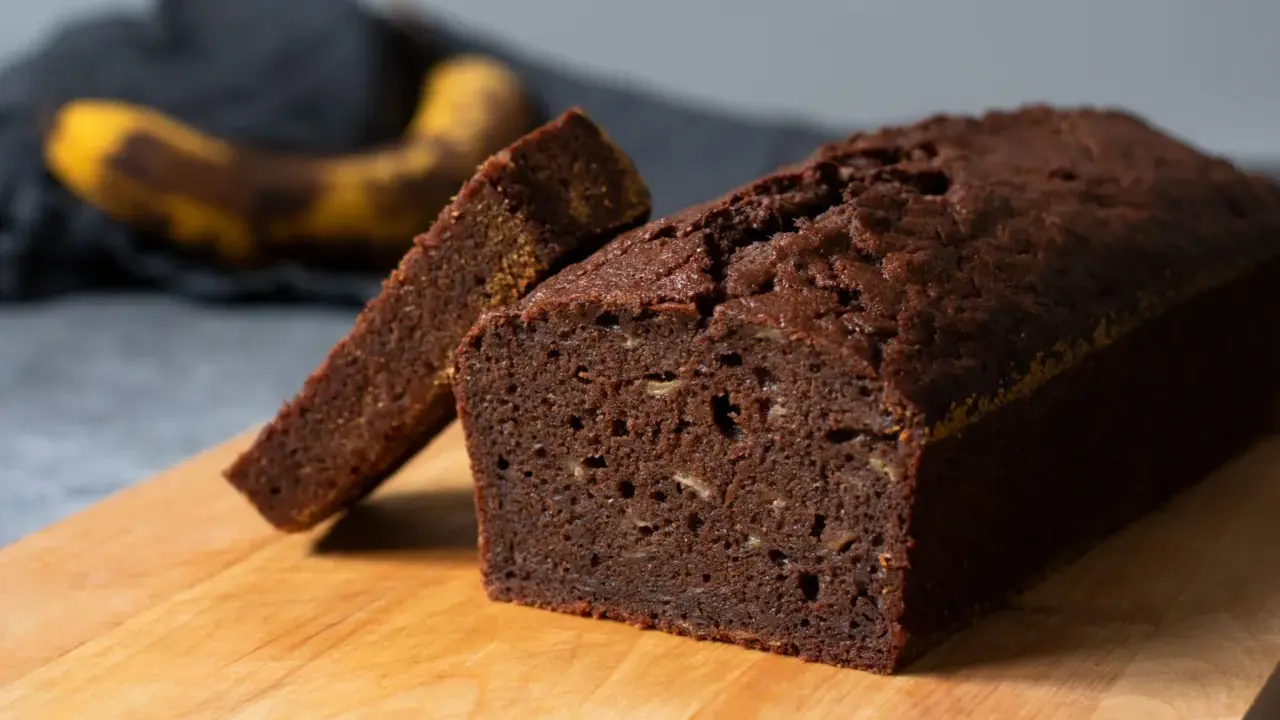 A slice of moist chocolate banana bread is propped against a whole loaf on a wooden cutting board.