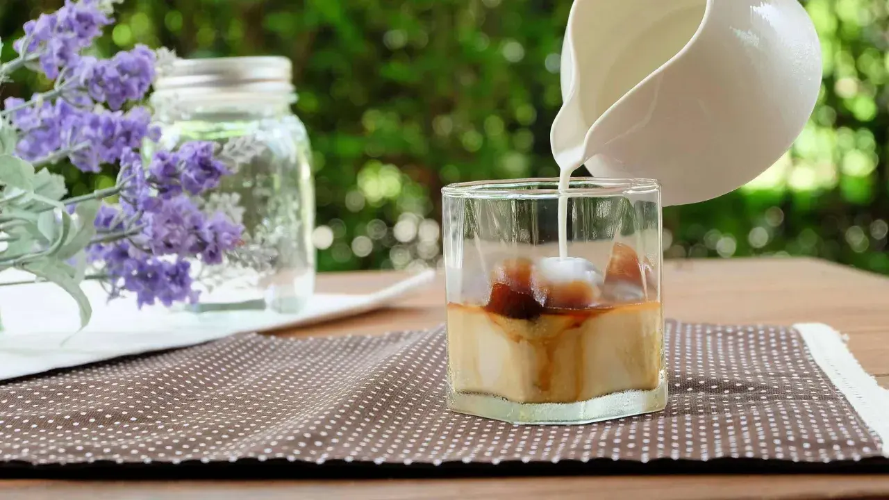 Milch wird in ein Glas mit Eiswürfeln und Kaffee gegossen, um einen erfrischenden Lavendel Latte zuzubereiten.