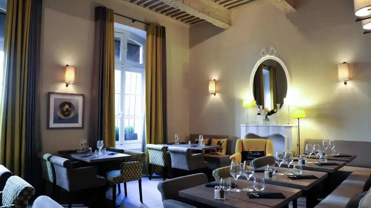 Salle à manger élégante avec tables dressées, prête à servir une spécialité de Provence. Lumière douce, rideaux ocre et gris, miroir orné.