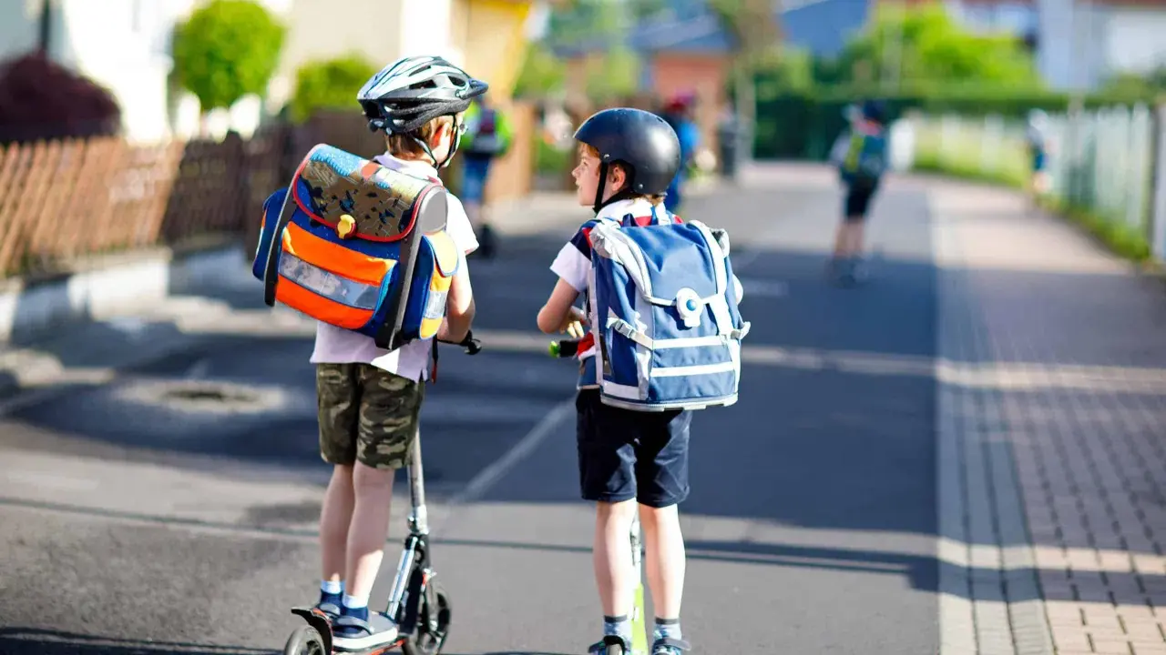 Deux enfants casqués, sacs à dos sur les épaules, se déplacent en trottinette. L'âge trottinette électrique est idéal pour ces jeunes écoliers.