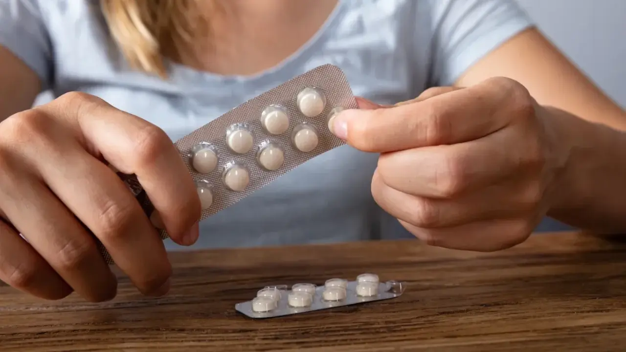 Dłonie trzymają opakowanie z tabletkami, które mogą być lekami z serotoniną. Na drewnianym blacie leży drugie, częściowo zużyte opakowanie.