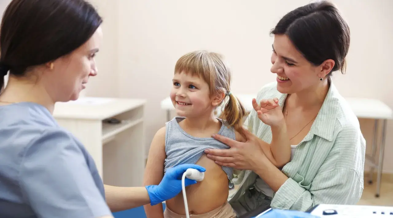 Lekarz wykonuje usg brzuszka niemowlaka. Uśmiechnięta dziewczynka i jej mama towarzyszą badaniu.