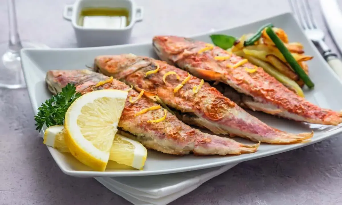 Cuatro salmonetes al horno, dorados y jugosos, servidos con rodajas de limón, perejil y patatas fritas.