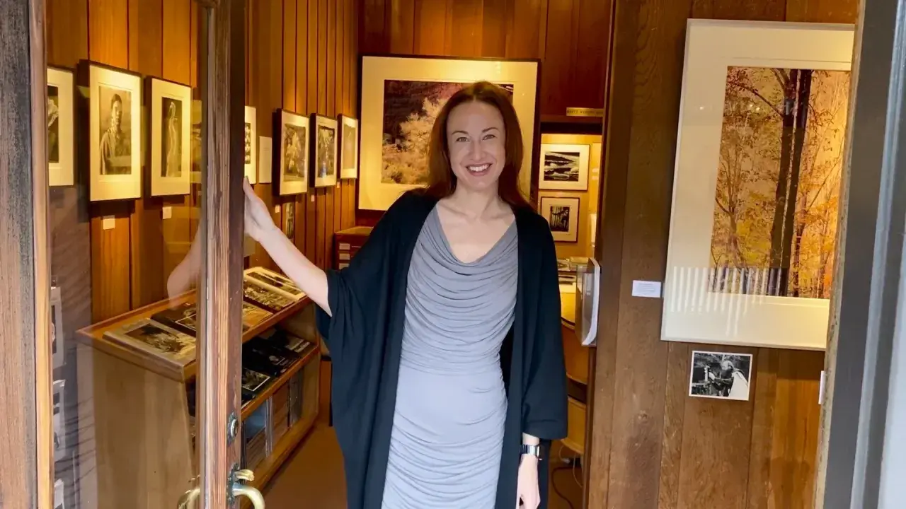 A woman smiles, opening the door to a photography west gallery filled with framed prints and display cases of artwork.