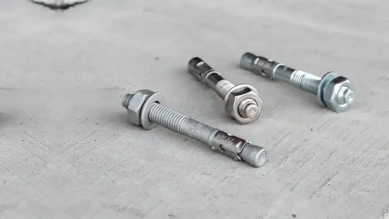 Trzy metalowe kotwy leżą na betonowej powierzchni. Każda kotwa ma gwintowany pręt i nakrętkę.