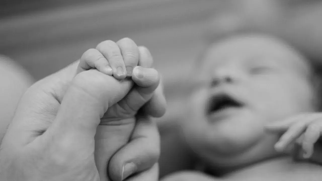 Dłoń dorosłego trzyma maleńką dłoń noworodka. Wiek postkoncepcyjny jest widoczny w delikatności paluszków.