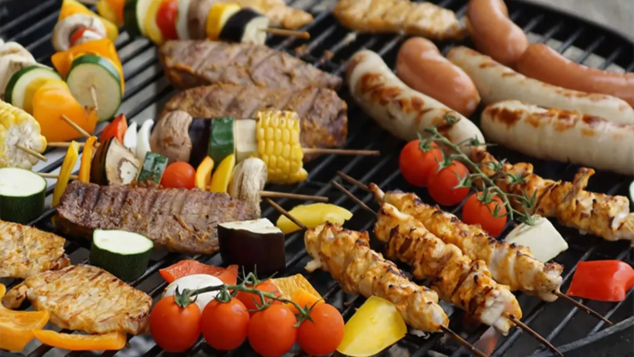 Una parrillada de que es bbq: brochetas de pollo y verduras, salchichas, bistecs y mazorcas de maíz asándose en una parrilla.