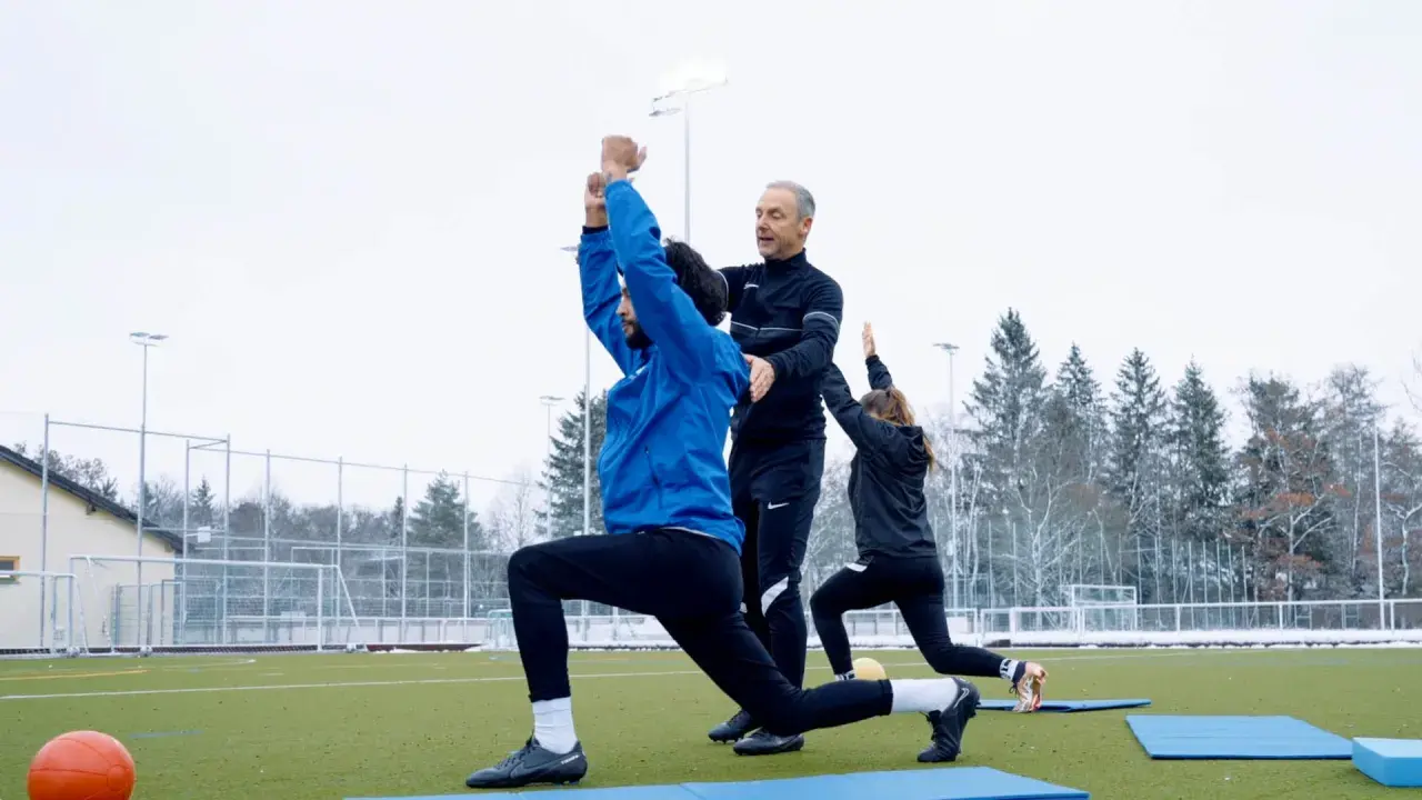 Athletik Training Fußball: Trainer korrigiert Ausfallschritt eines Spielers auf Kunstrasen. Zwei weitere Spieler machen Übungen im Hintergrund.