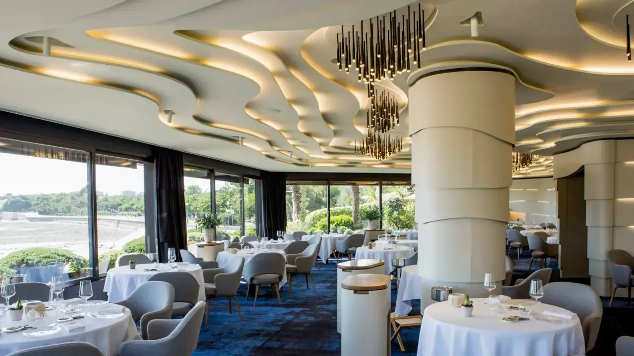 Salle élégante d'un restaurant chef de baie, tables dressées, vue sur la mer et jardin luxuriant. Plafond ondulé et luminaires modernes.