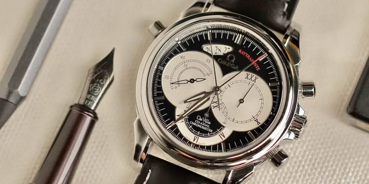 Elegante cronografo rattrapante Omega De Ville con quadrante nero e contatori argentati, accanto a una penna stilografica.