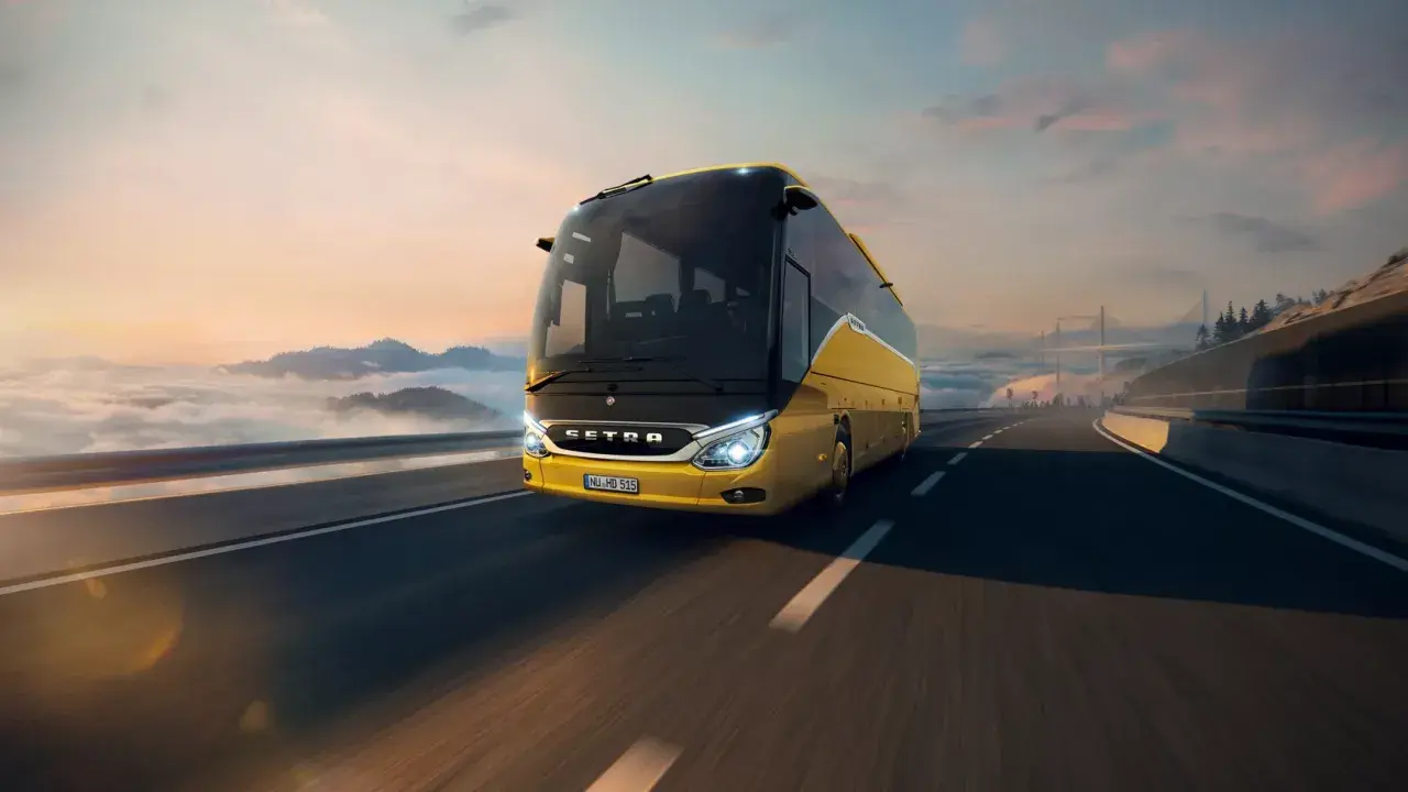 Autobús Setra amarillo brillante viaja por una carretera escénica al amanecer, con nubes y montañas de fondo.