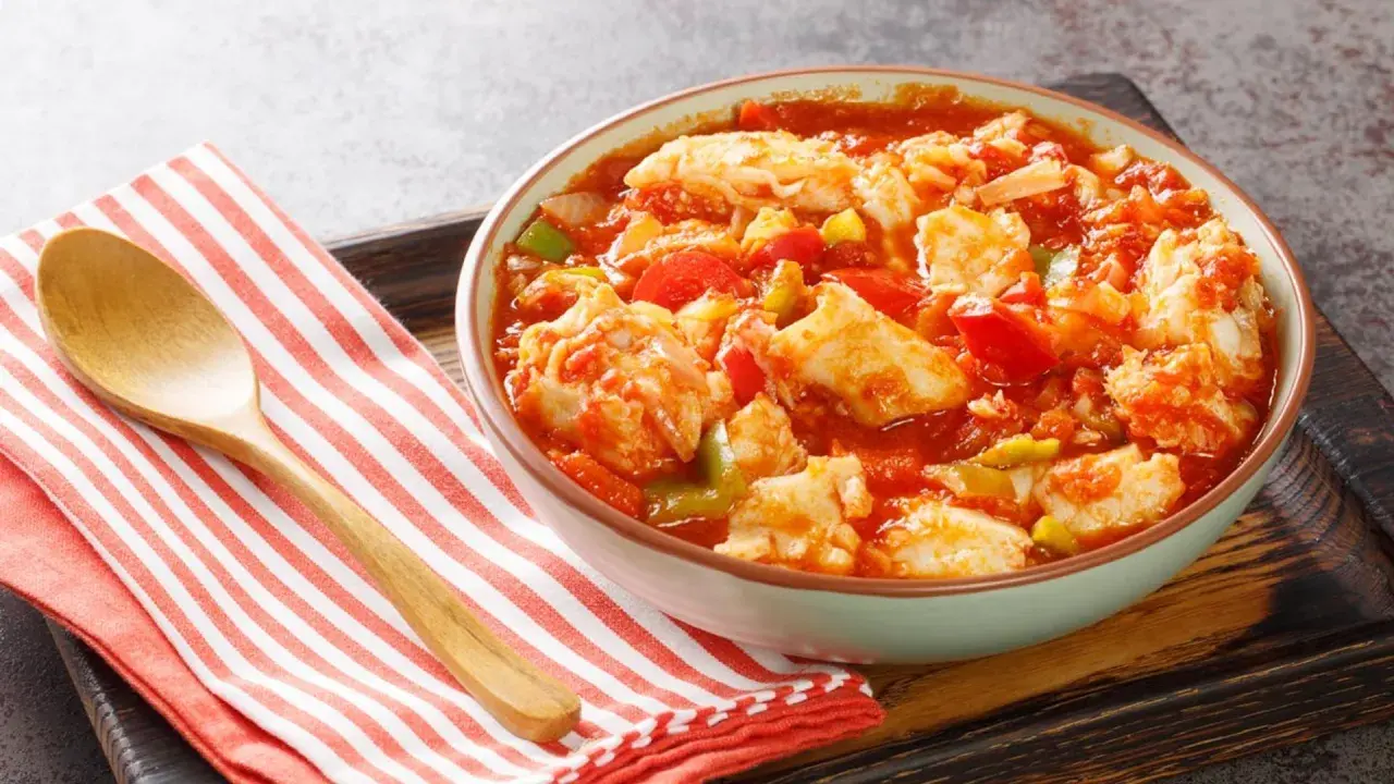 Un tazón de bacalao ajoarriero con pimientos y tomate, servido con una cuchara de madera sobre una servilleta a rayas.