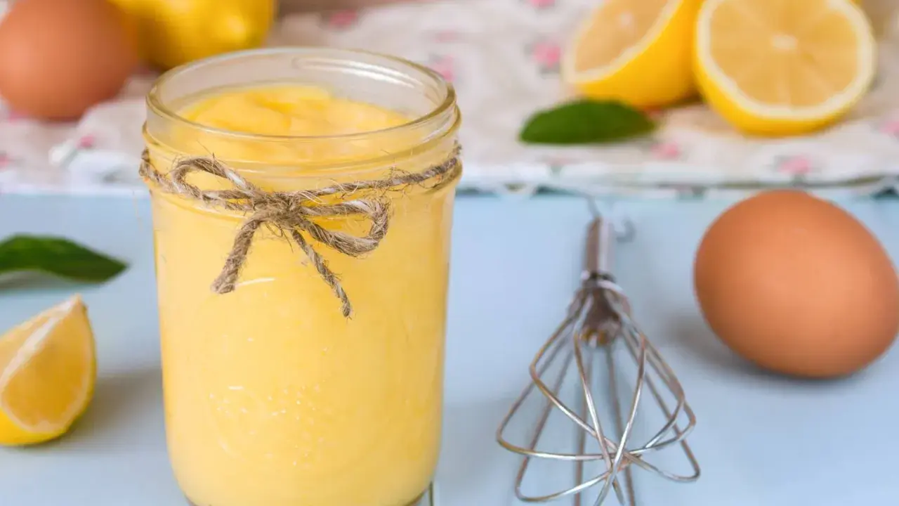 Un tarro de lemon curd casero, atado con cuerda, junto a limones, huevos y un batidor.