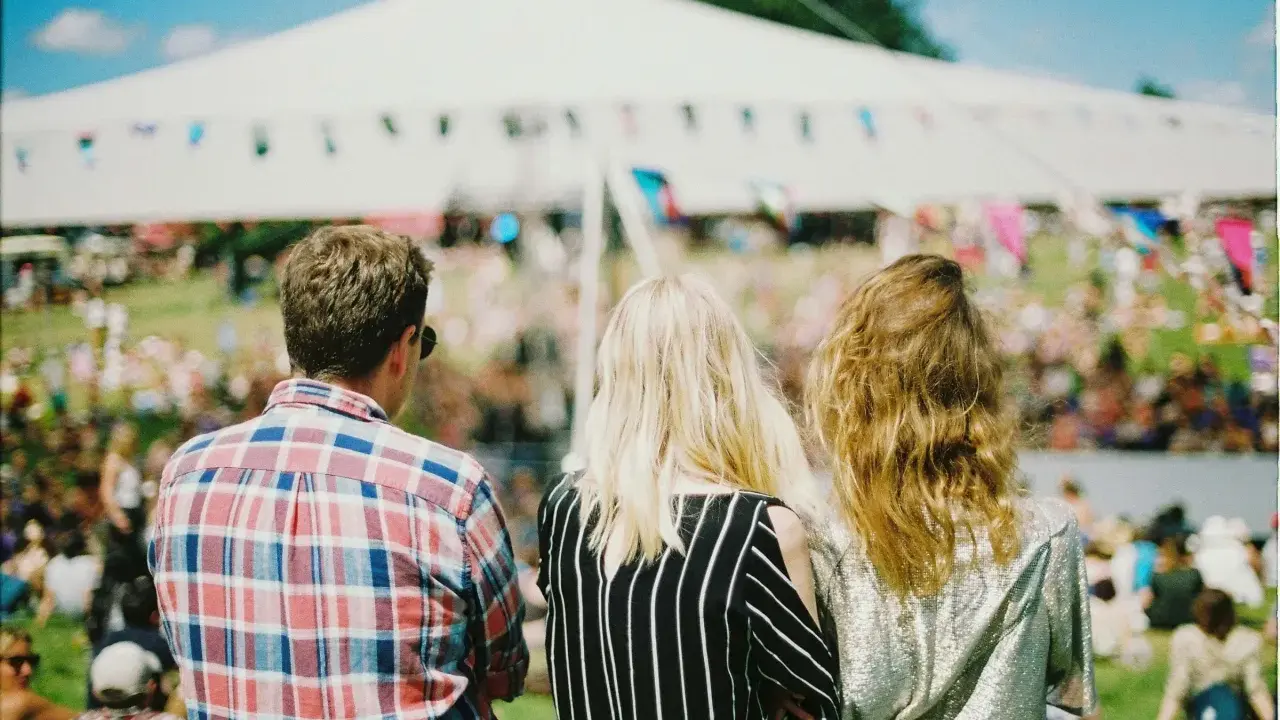 Tre vänner njuter av en solig dag på en musikfestival i Stockholm. En stor vit tältduk och flaggor syns i bakgrunden.
