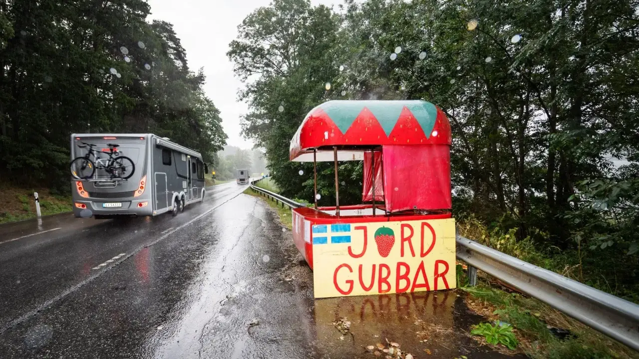 Regnig dag vid vägkanten med en jordgubbsförsäljning. Husbilen passerar, kanske på väg till sommarstugan under fruntimmersveckan, som bondepraktikan säger.