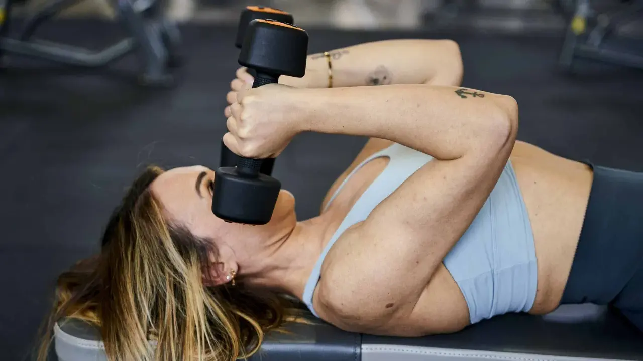 Donna sdraiata esegue skullcrushers con manubri, un esercizio efficace per i gruppi muscolari da allenare insieme, come tricipiti e spalle.