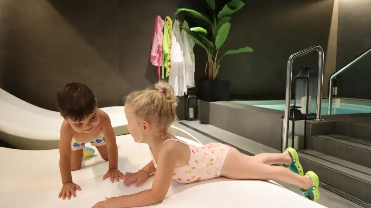 Niños jugando en un spa para niños, con una piscina al fondo y toallas colgadas.