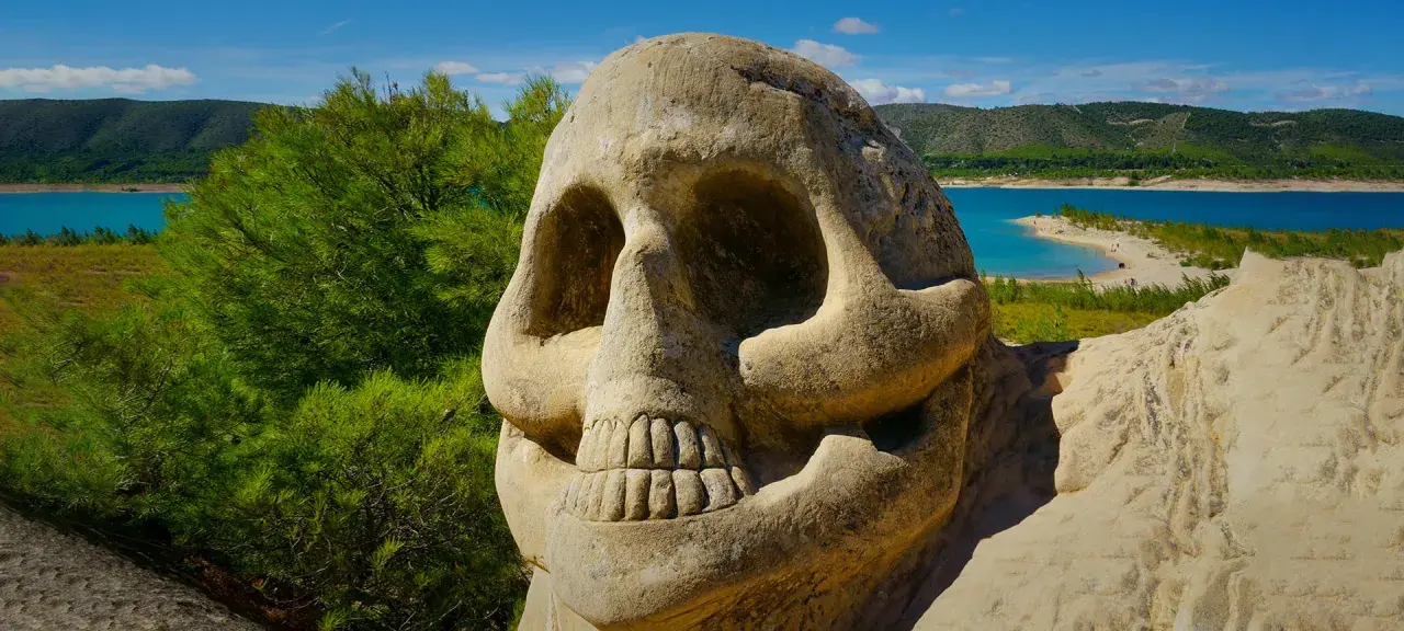 Escultura de calavera en la **ruta de las caras**, con un lago azul y colinas verdes al fondo.