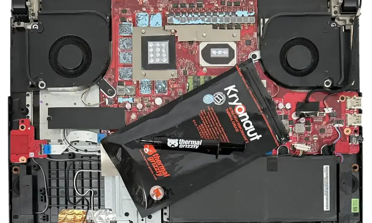 Wnętrze laptopa z wentylatorami i płytą główną, gotowe do wymiany pasty termoprzewodzącej. Opakowanie Kryonaut Thermal Grizzly leży na komponentach.