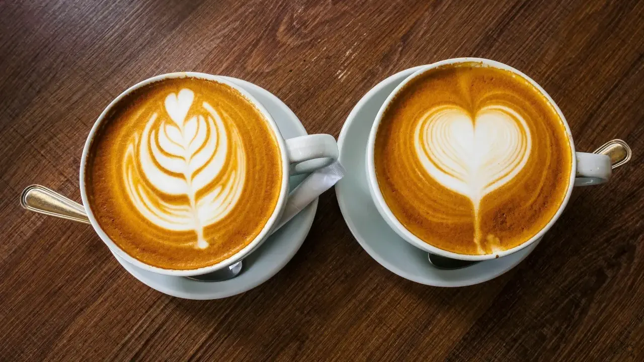 Zwei Cappuccinos mit Latte Art auf einem Holztisch. Sie bestehen aus Espresso, heißer Milch und Milchschaum.