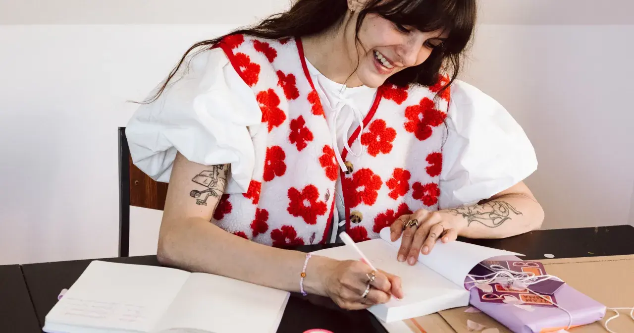Une jeune femme souriante fait une dédicace livre, stylo à la main, devant un cahier ouvert et un paquet cadeau violet.