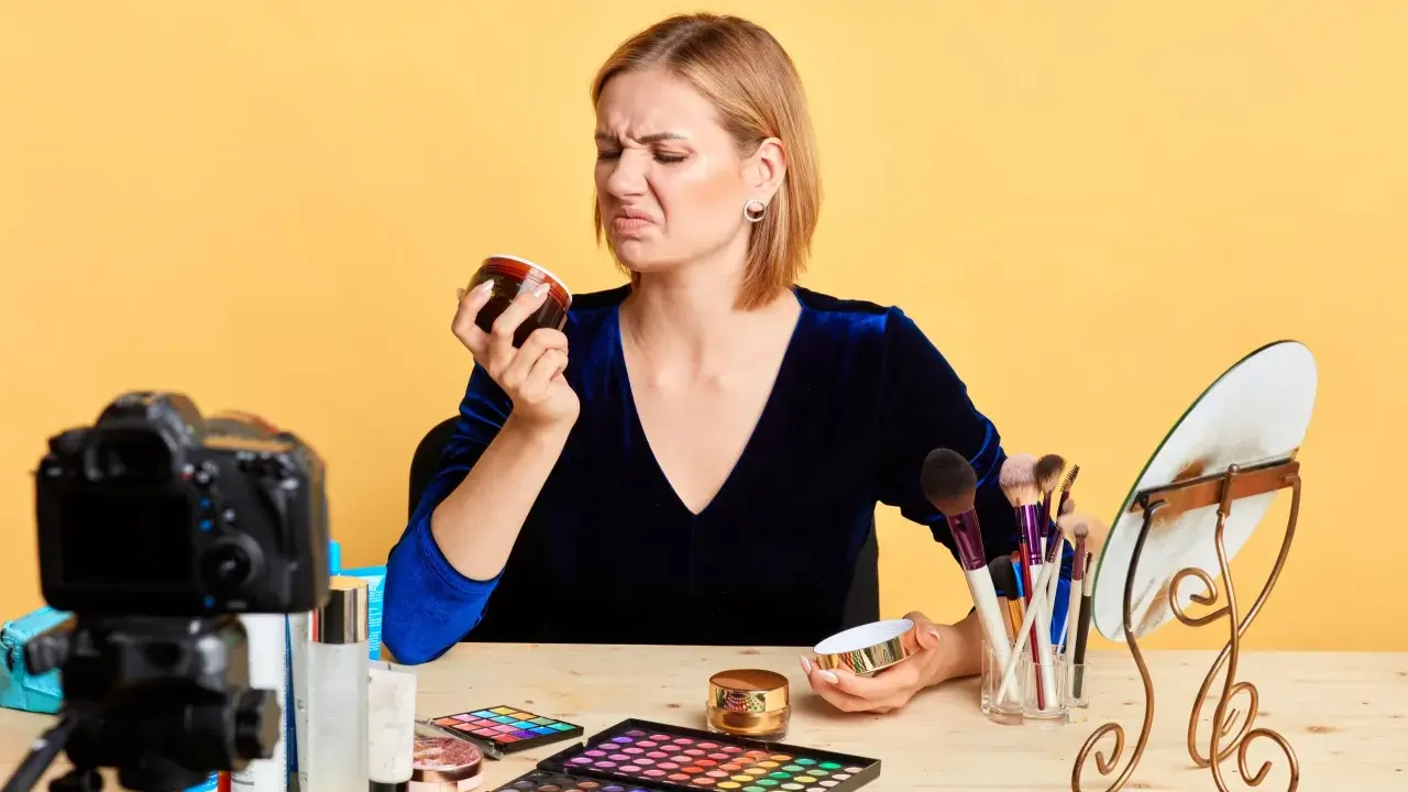 Donna con espressione disgustata annusa una crema viso chiusa, forse per verificarne la scadenza. Sul tavolo, trucchi e una telecamera.
