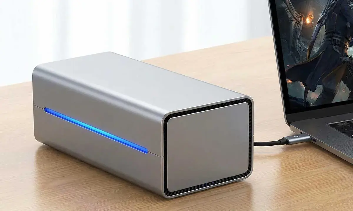 Srebrna obudowa egpu do laptopa z niebieskim podświetleniem, podłączona do laptopa z grą na ekranie.