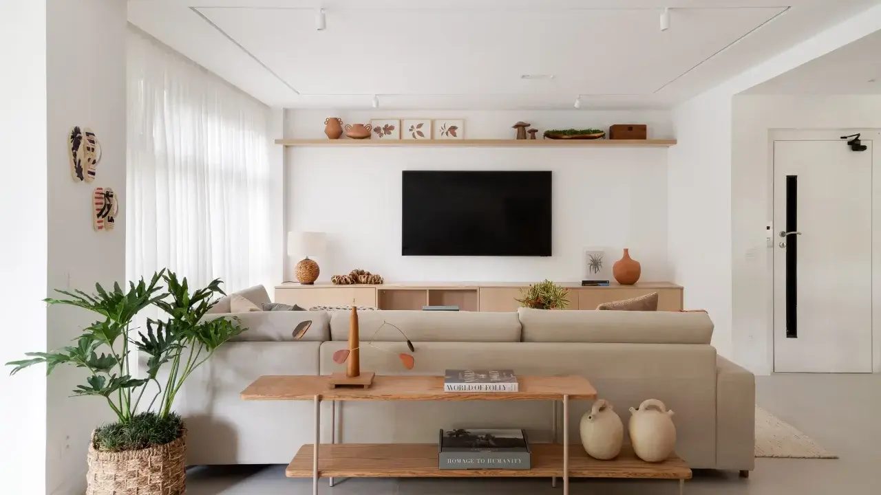 Estantería de madera con objetos decorativos, jarrones y cuadros. Un sofá beige y una planta grande complementan el espacio. Ideas de qué poner detrás del sofá.