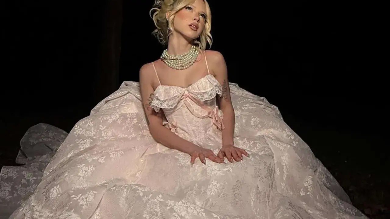 A woman in a voluminous, lace-trimmed gown and pearl necklace embodies modern romantic style, posing in a dark, outdoor setting.