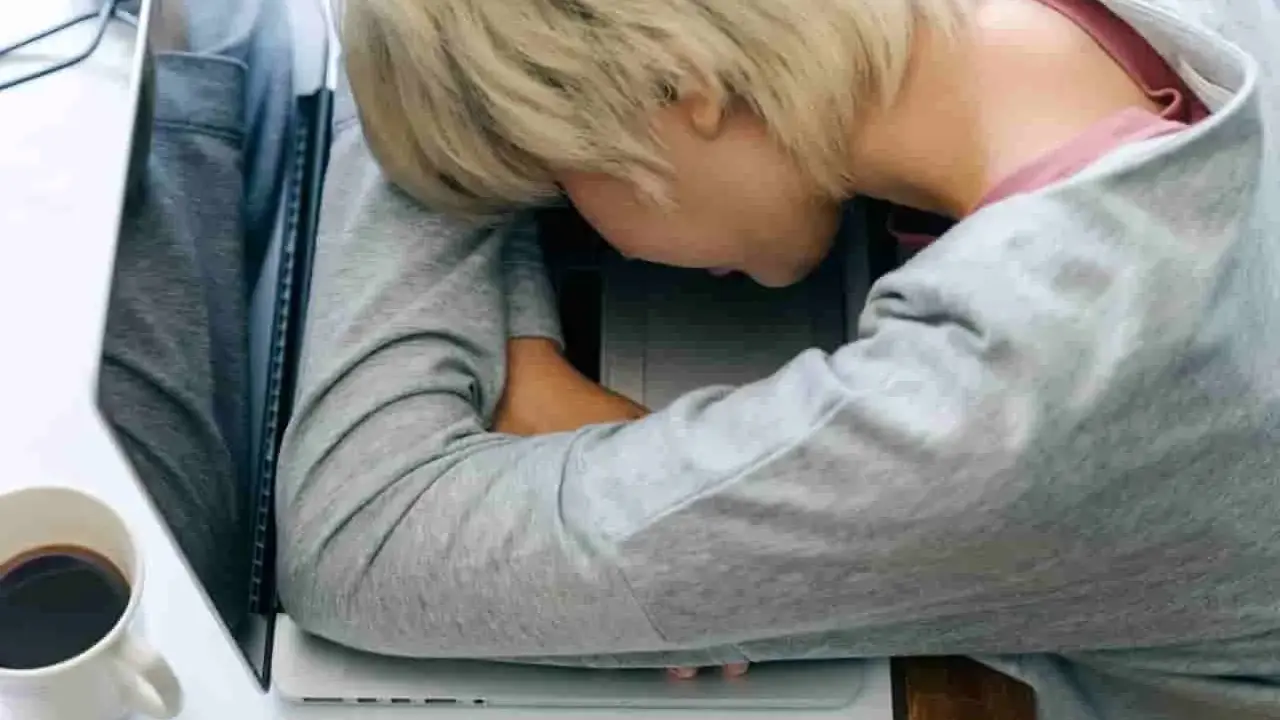 Zmęczona osoba śpi na laptopie. Czy leczenie odwykowe a zwolnienie z pracy to trudny wybór? Kawa obok.