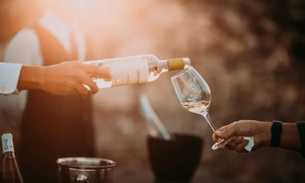 En person häller upp ett glas vin från en flaska. Solen skiner och skapar en varm atmosfär.