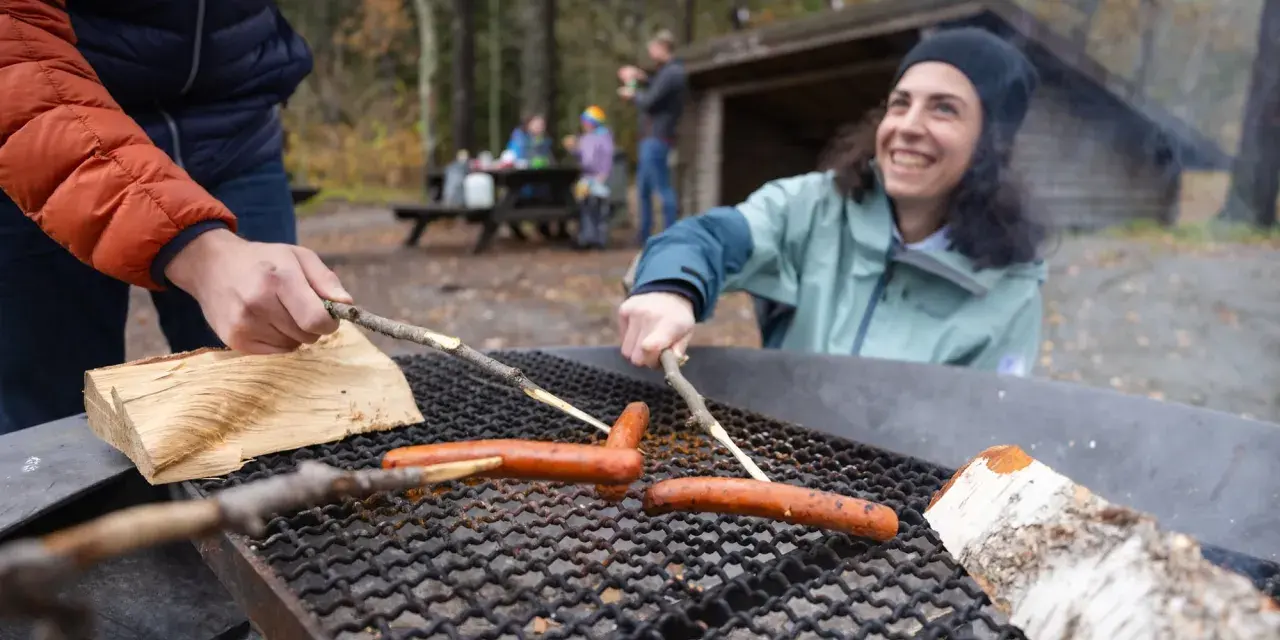 Vänner grillar korv vid en eldstad på Otto Pers gård. En kvinna ler glatt medan hon grillar.