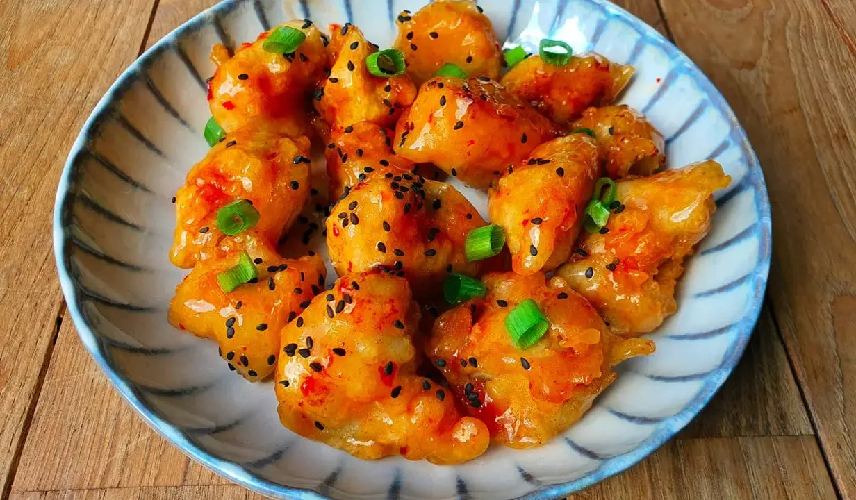 Krispiga bitar av sweet chili kyckling, toppade med sesamfrön och salladslök, serveras på en blårandig tallrik.